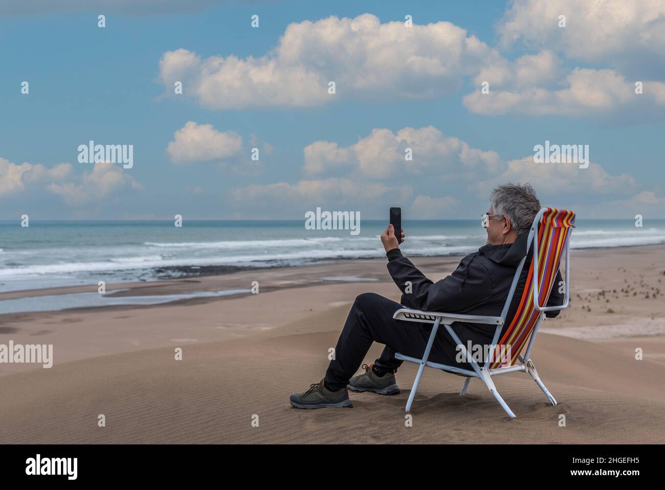 Mature adult male sitting on a beach chair on top of a dune facing the sea talking on video with smartphone. Horizontal Stock Photo