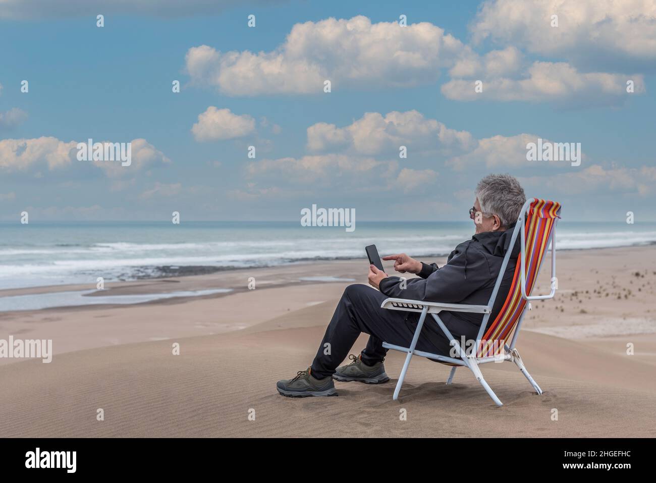 Adult man sitting on a beach chair on top of a beachfront medano typing on smartphone. Horizontal photo Stock Photo