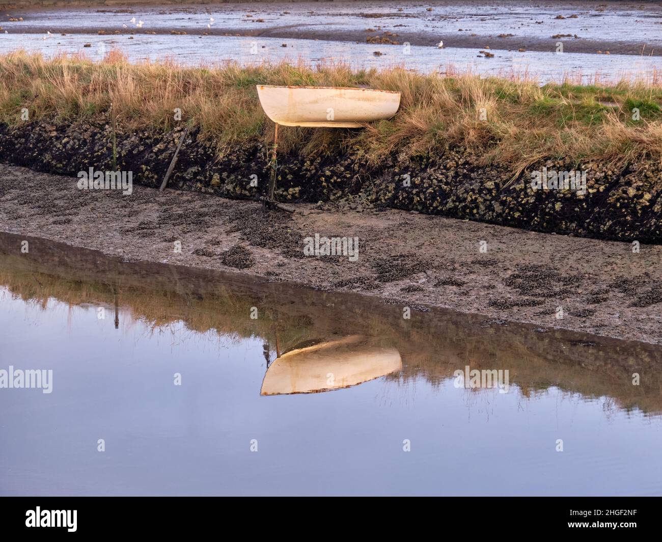 A small rowing boat is left high and dry when the tide goes out.  It is supported at the stern by the bank and at the bow by its mooring post. Stock Photo