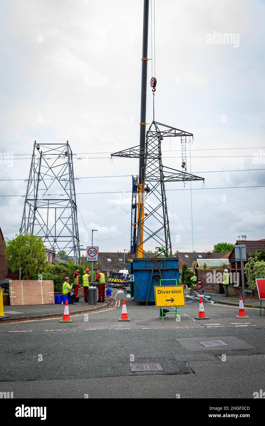 Engineers and Technicians watch while the upper half of an electricity pylon is removed and lowered to the ground by crane Stock Photo