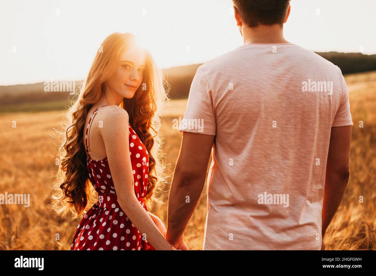 Redheaded girl holding her boyfriend's hand walking through wheat field holding hands. For lifestyle design. Holding hand. Wheat field. Stock Photo