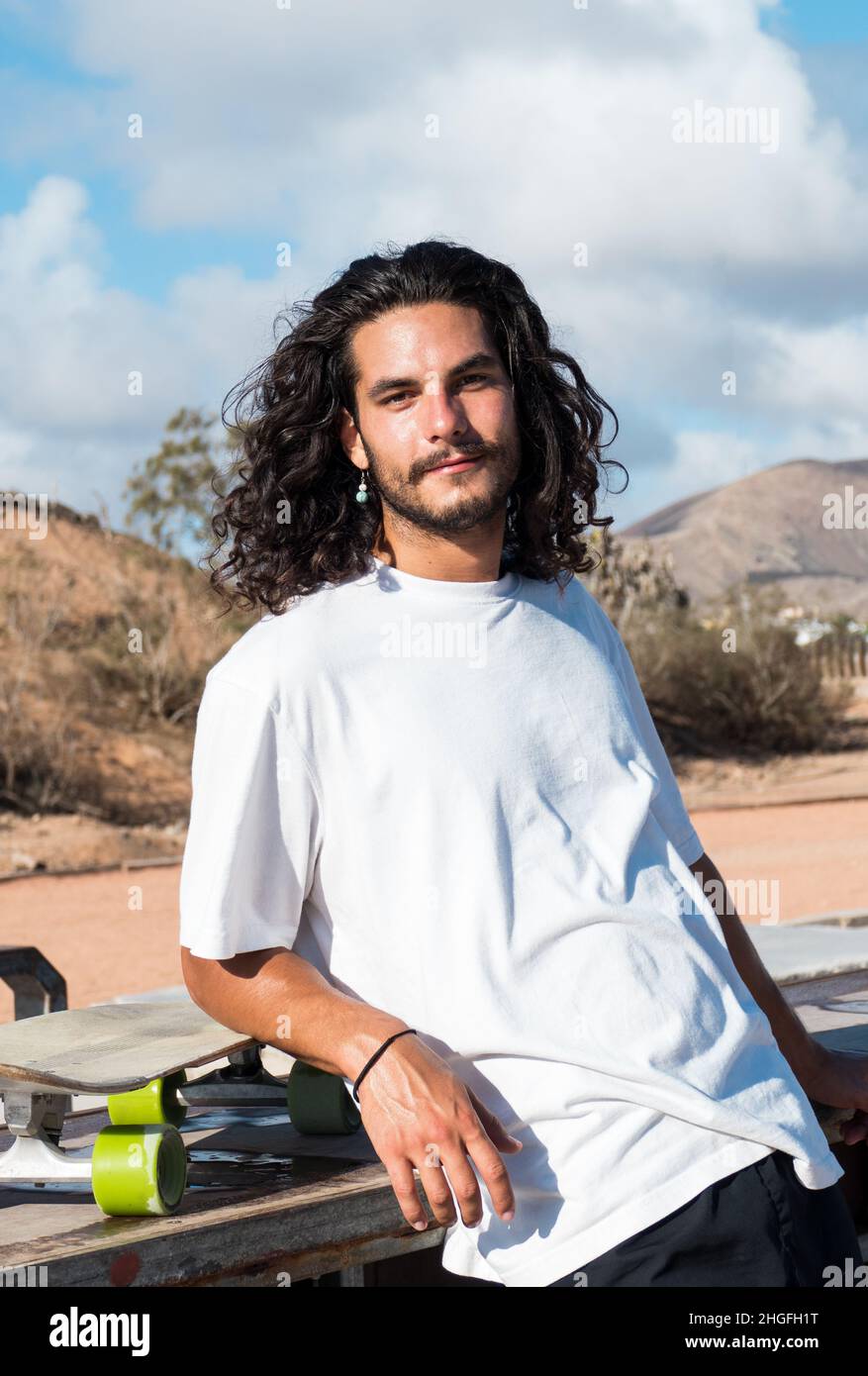 Young caucasian skater portrait. He has long black hair and he has a beard. He is wearing white t shirt. Stock Photo