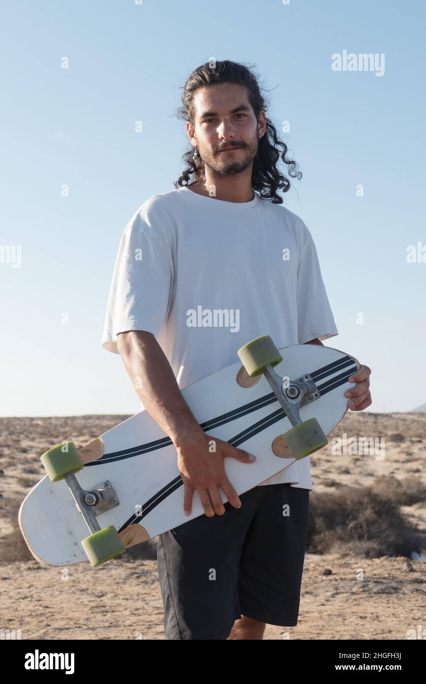 Young adult skater portrait standing outdoors holding his skateboard. He has long black hair and a beard. Stock Photo