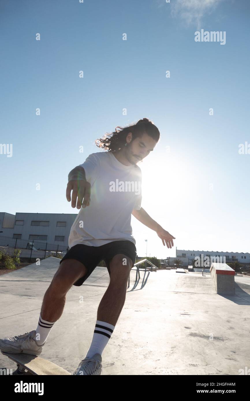 Young male caucasian skater at the skate park ramp. He is wearing summer clothes and he has long black hair and a beard. Stock Photo