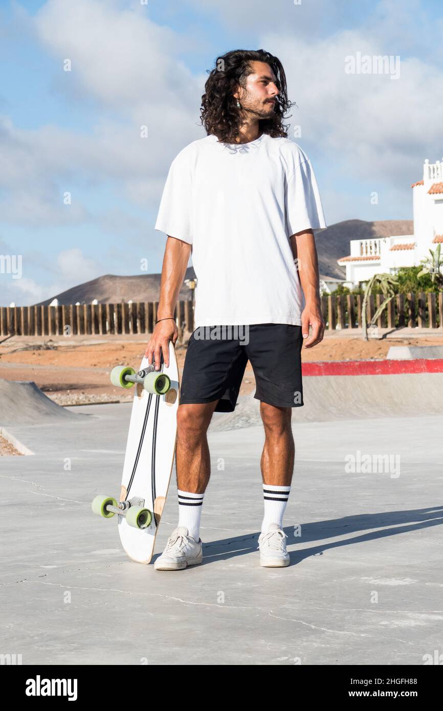 Young male caucasian skater standing at the skate park. He is holding his surfskate skateboard and whearing summer clothes. He has long black hair. Stock Photo