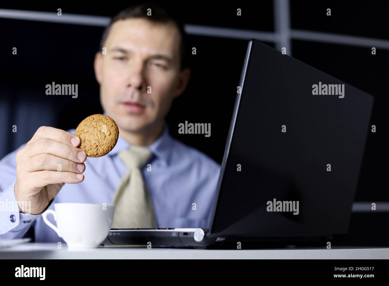 Man eating oatmeal cookie sitting at laptop. Comfort food, sweet dessert against stress, break during work in office Stock Photo