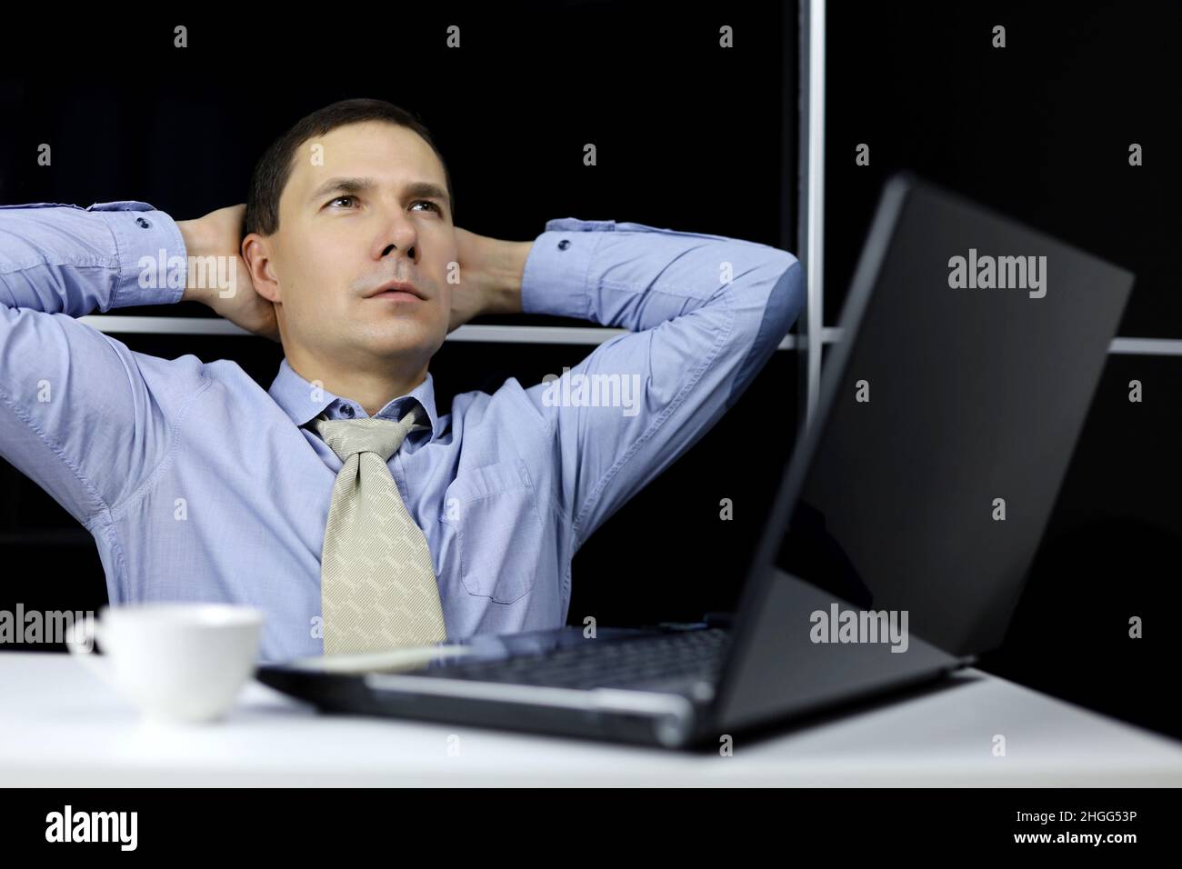 Man in blue shirt and tie resting with his hands behind his head sitting at laptop. Break in office, dreaming at work, inspiration and reflections Stock Photo