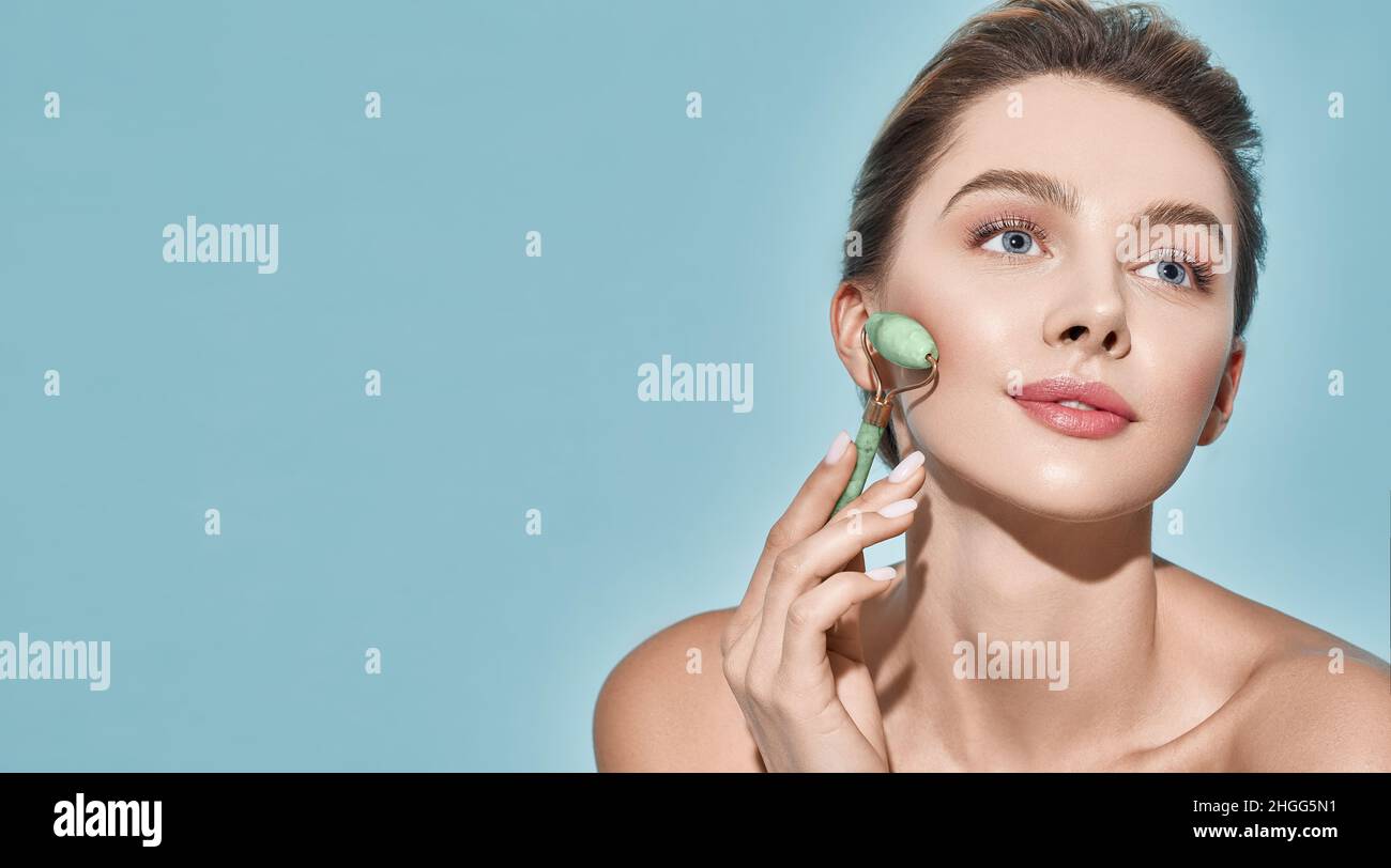 Beautiful woman using stone facial roller for elastic perfect skin, isolated on blue background. Jade roller with natural quartz stones at cosmetology Stock Photo