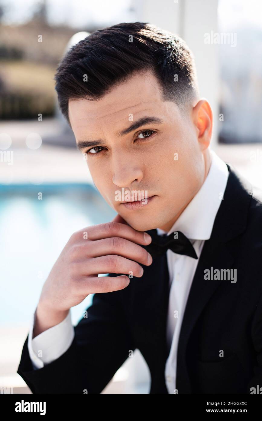Portrait of handsome caucasian groom with dark hair and brown eyes looking  at camera while posing outdoors. Young man in stylish black suit sitting on  Stock Photo - Alamy