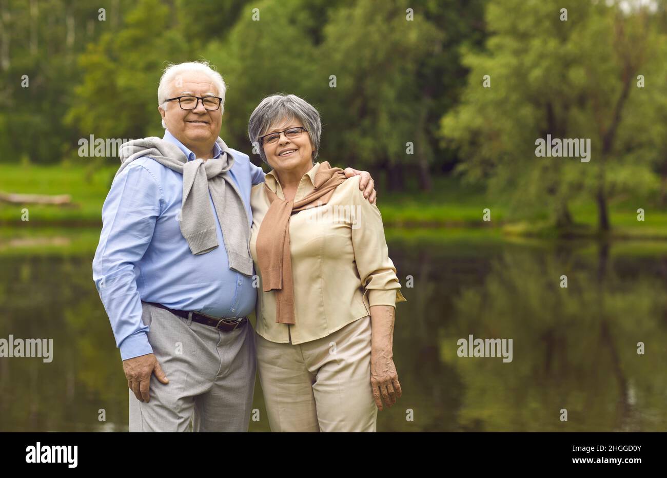 Portrait of smiling elderly pensioners standing in the park by the lake in summer. Stock Photo