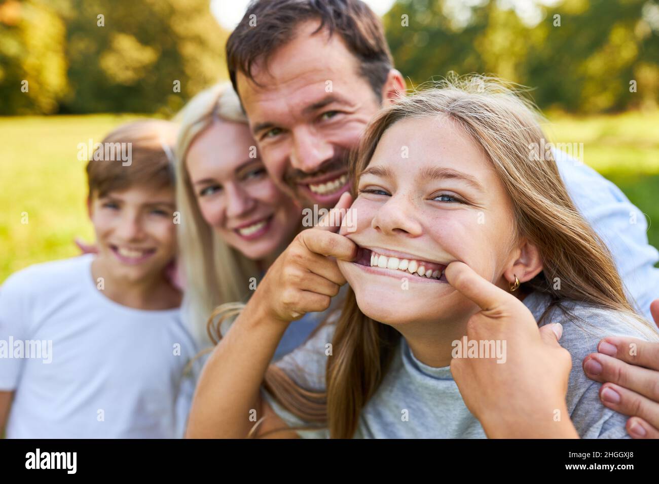 Girl makes a funny grimace with brother and parents in the background in the park Stock Photo