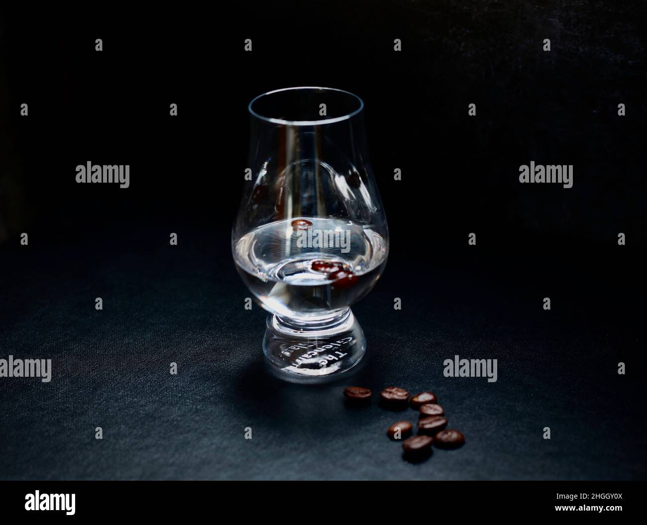 Chiaroscuro image of Sambuca in a simple glass with coffee beans . Dark background with space around and a handful of coffee beans in the foreground. Stock Photo