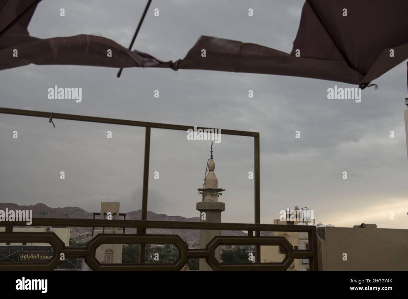 View of a mosque in Aqaba Jordan with grayish blue skies at the Red Sea through a torn umbrella awning at sunrise. Stock Photo