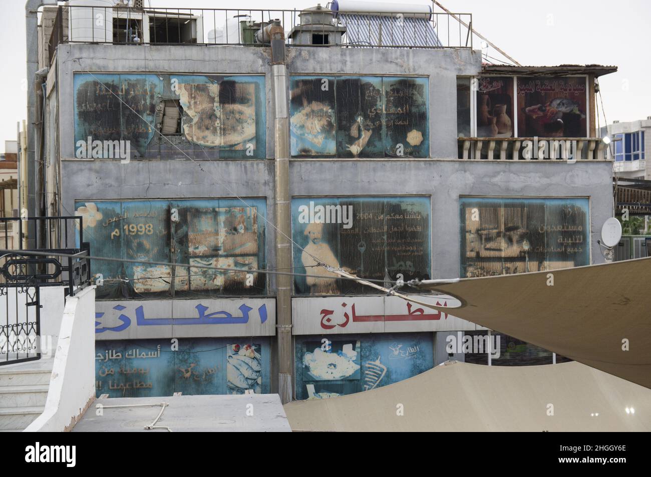 Faded and windswept billboards on a building in Aqaba Jordan depicting bread making and a restaurant and Arabic writing and torn awnings. Stock Photo