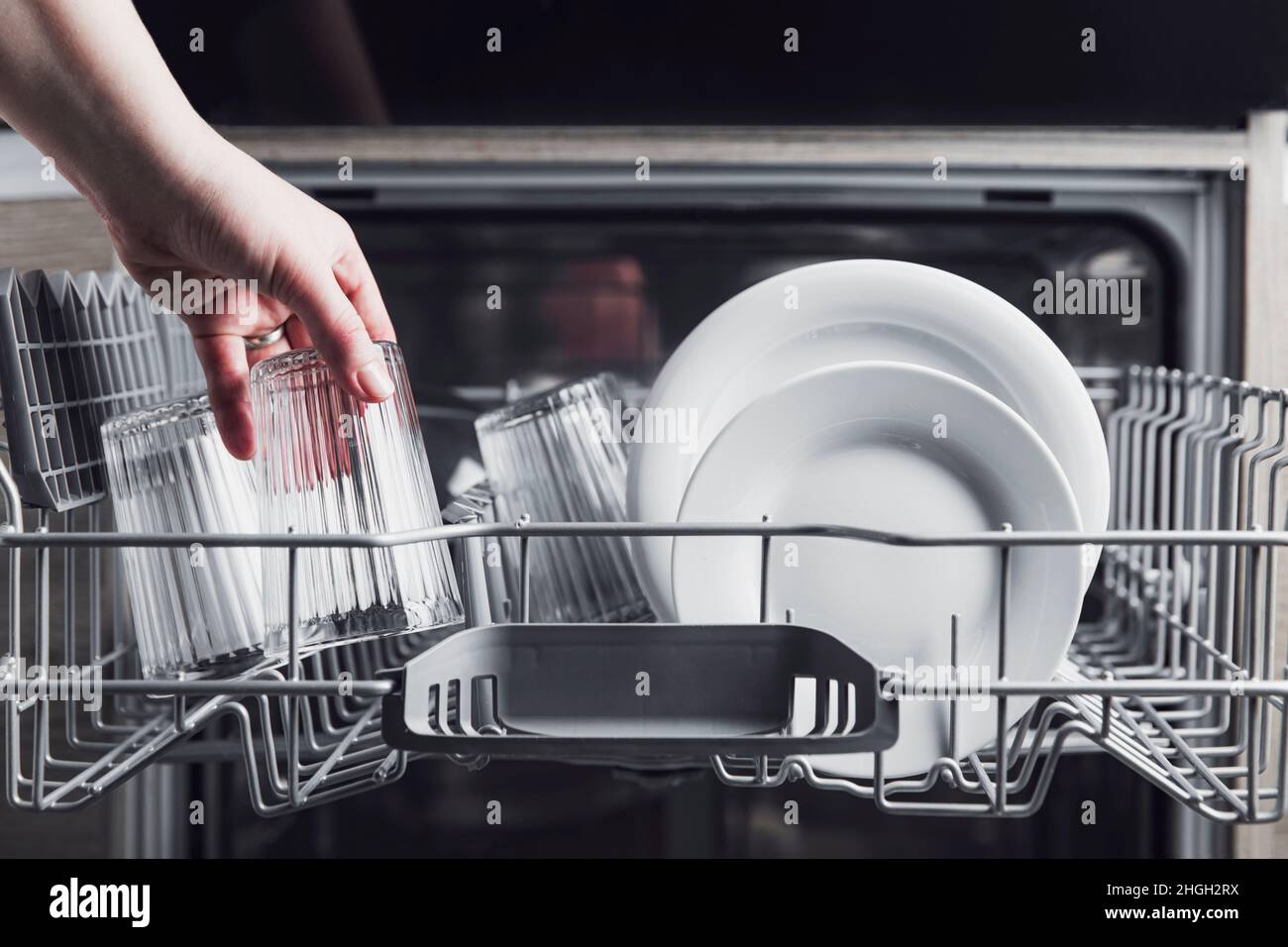Female hand loading dished, empty out or unloading dishwasher with utensils. Kitchen appliances, lifestyle view. Woman puts a plate in the dishwasher or takes from it. Housewife does her housework  Stock Photo