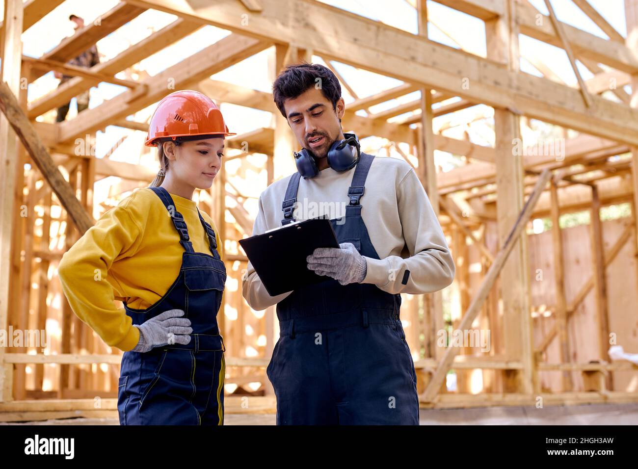 Two man and woman in uniform and helmet explore construction documentation on the building site near the wooden building constructions, have conversat Stock Photo