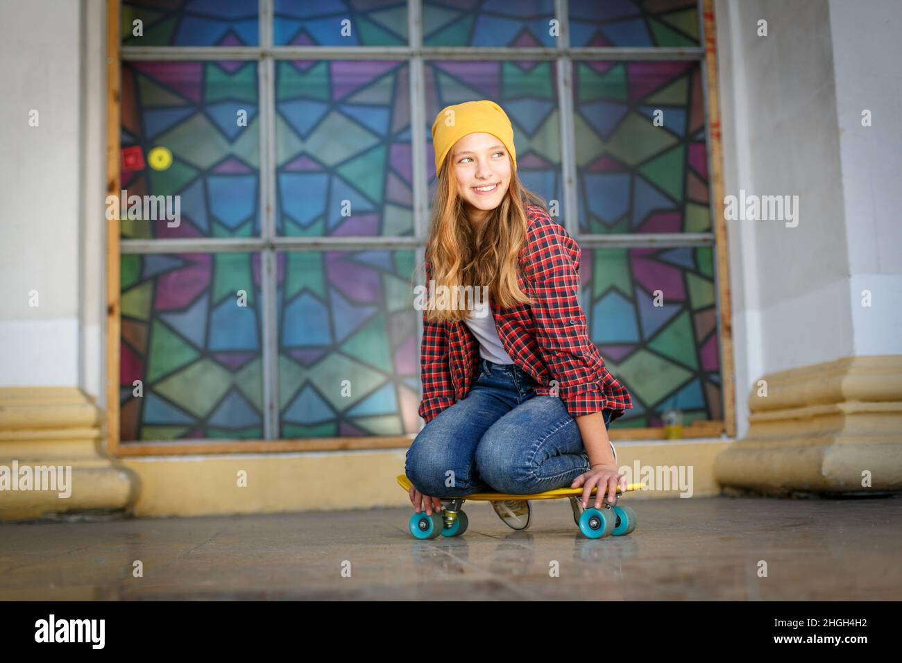 Lifestyle horizontal outdoor portrait of young smiling teenage girl  with a yellow skateboard wearing yellow hat and red plaid shirt Stock Photo