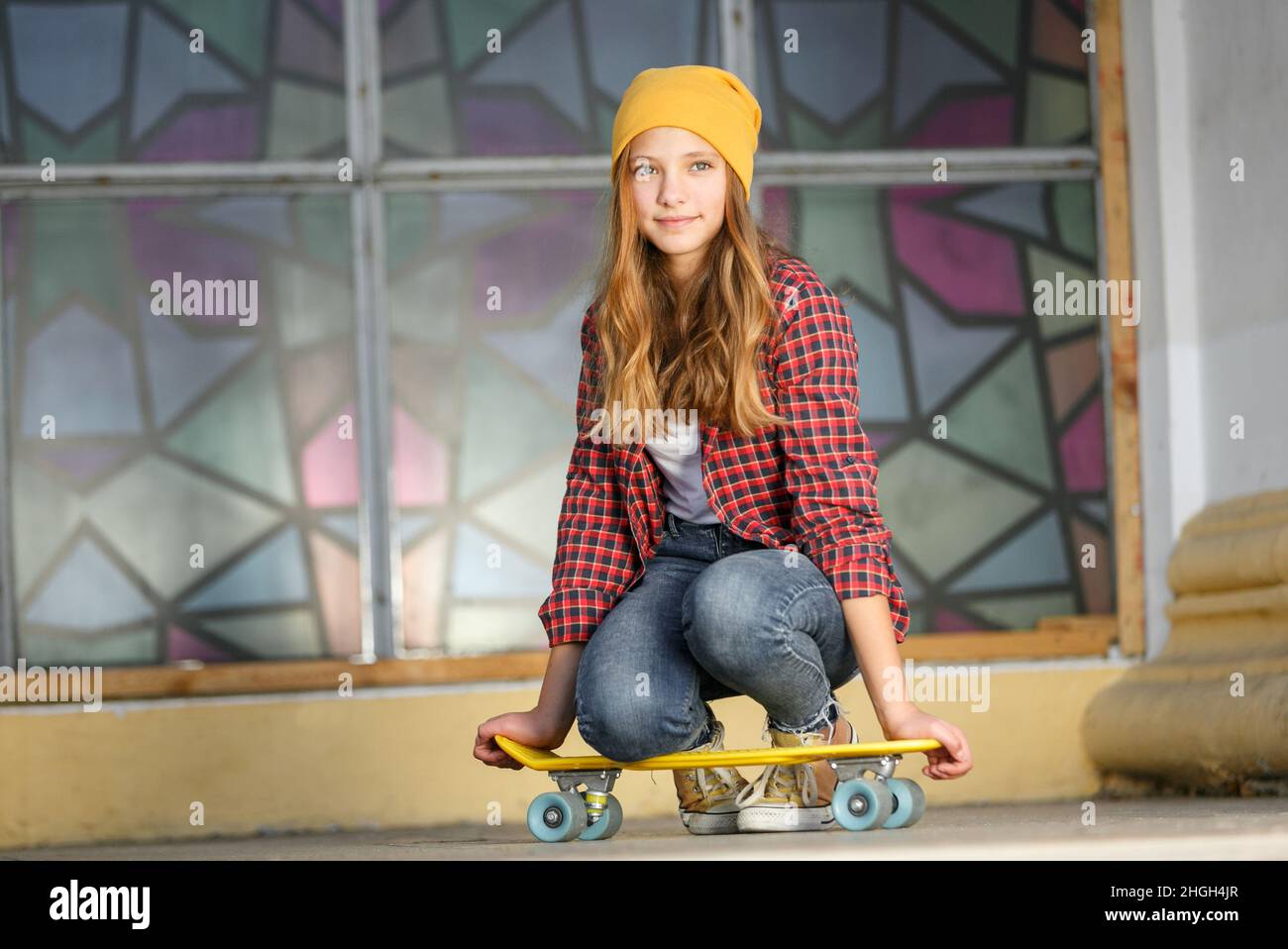 Lifestyle horizontal outdoor portrait of young smiling teenage girl  with a yellow skateboard wearing yellow hat and red plaid shirt Stock Photo