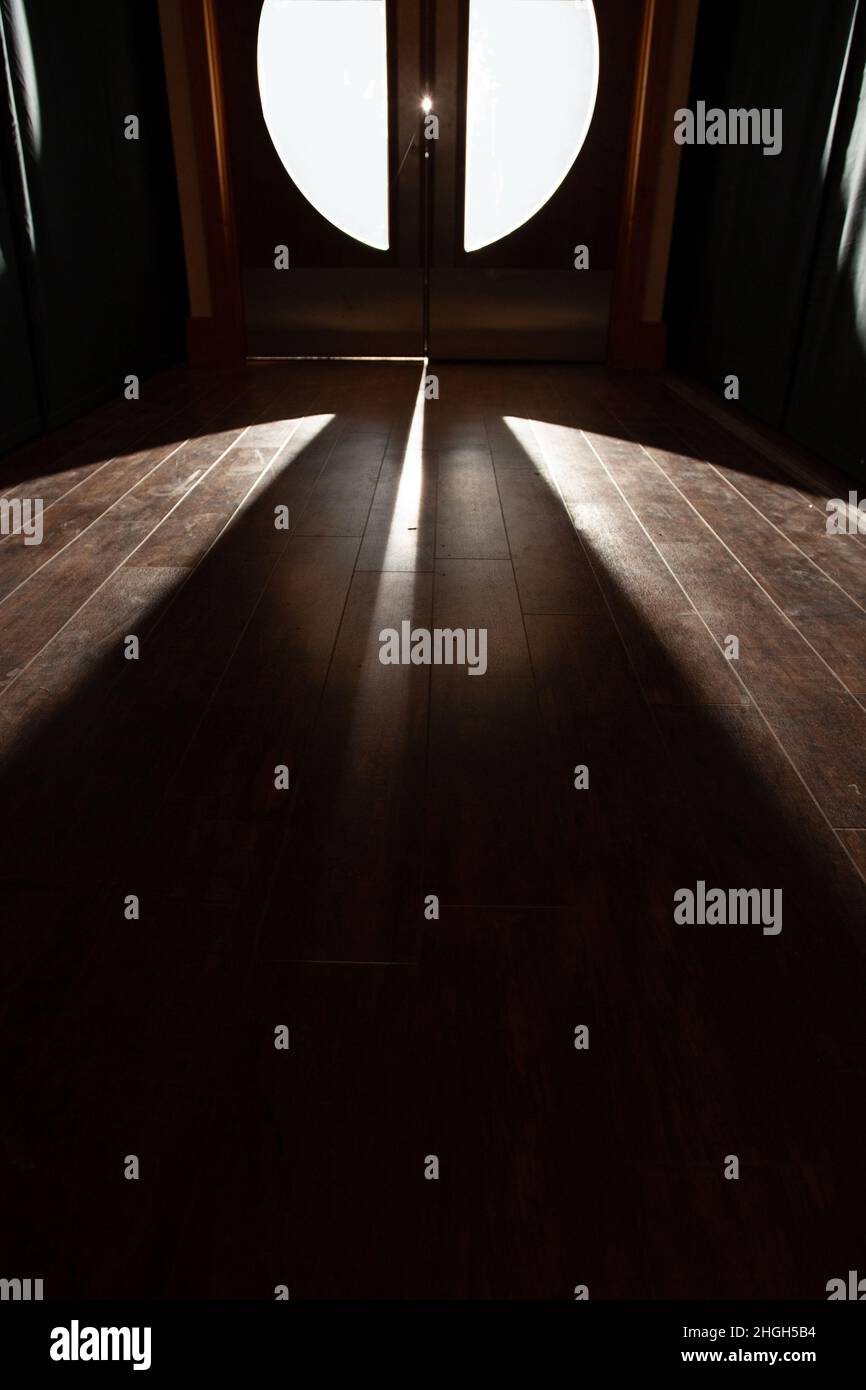 An Erie spooky light coming through a set of glass double doors shining onto a wooden floor with a corridor effect. Conceptual image various meanings. Stock Photo