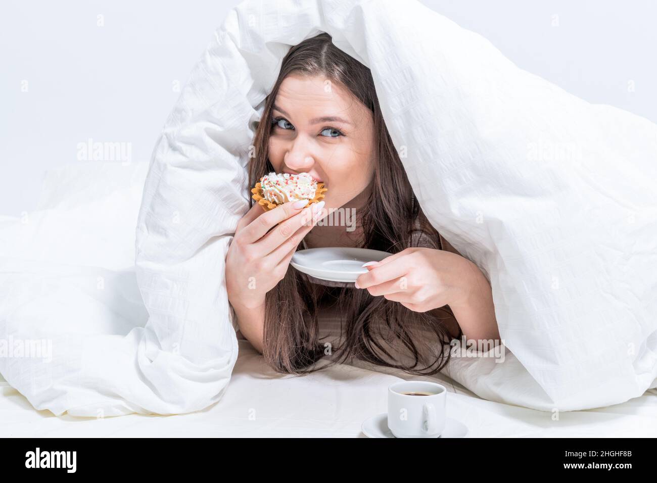 Cute woman is eating a cupcake while lying on the bed, under the covers. Breakfast in bed. Dessert. Calories. Stock Photo