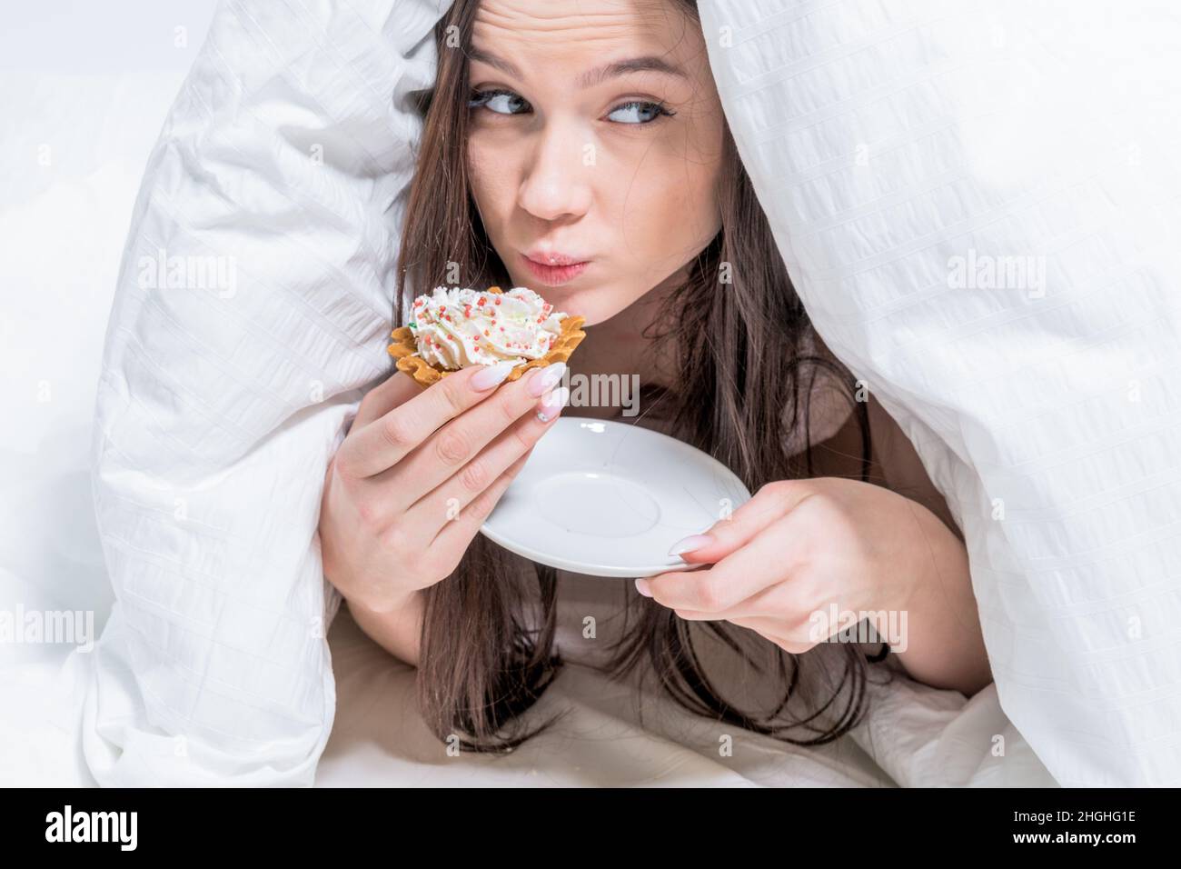 A cute woman is furtively, unnoticed, eating a cupcake, lying on the bed under the covers. Breakfast in bed. Dessert. Calories. Stock Photo