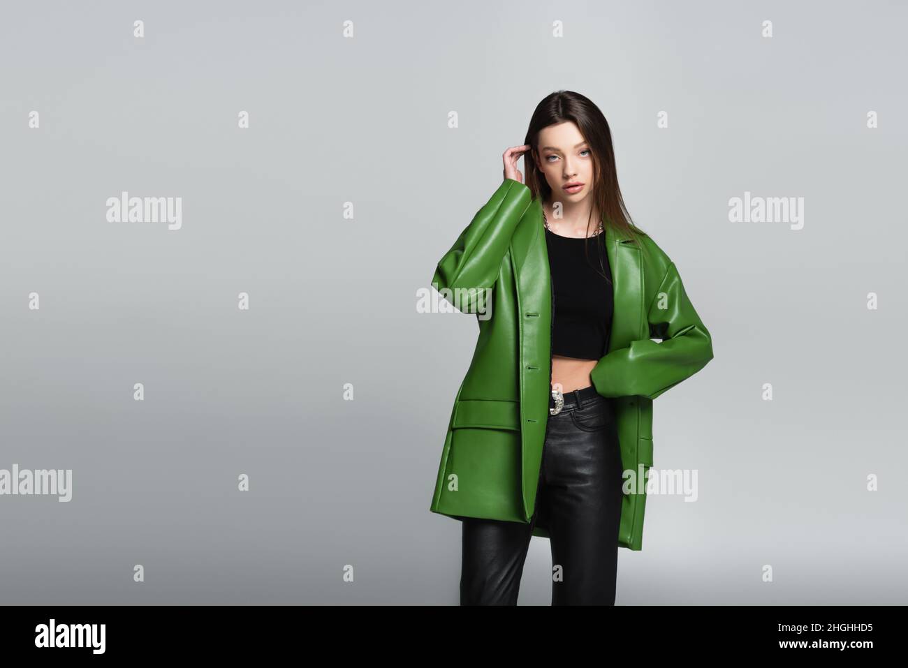 pretty woman in green jacket and black trousers standing with hand on waist isolated on grey Stock Photo