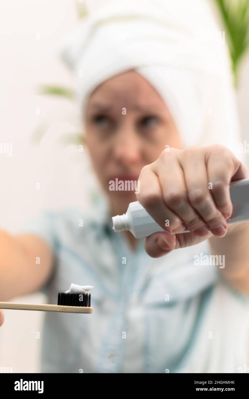 A young pretty woman with a towel on her head in a bright bathroom spreads toothpaste on a toothbrush against the background of a green plant. Selecti Stock Photo