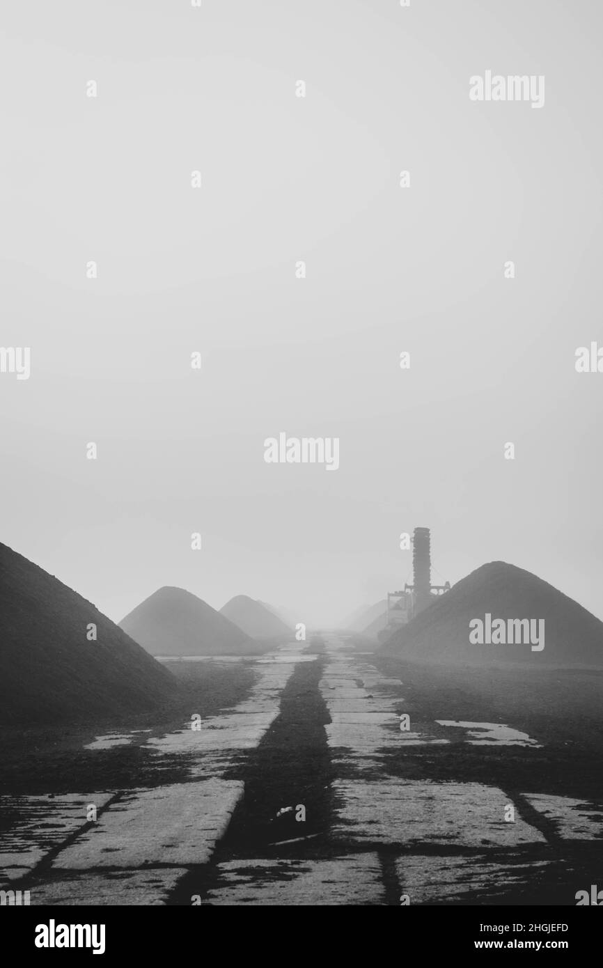 Black and white photo of concrete path leading through piles of peat on a foggy morning. Stock Photo