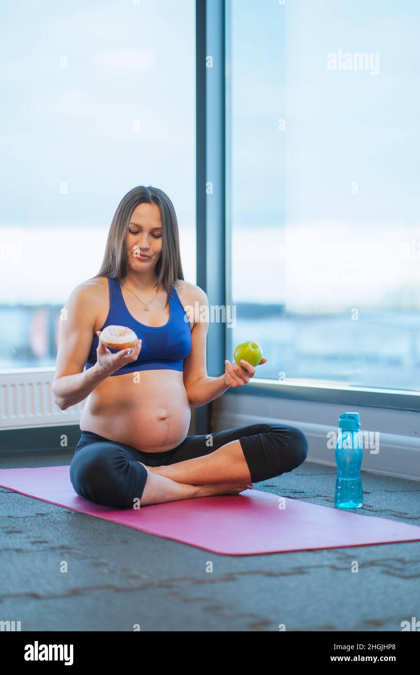 athletic pregnant woman in sportswear hold donut and apple sitting on yoga mat Stock Photo