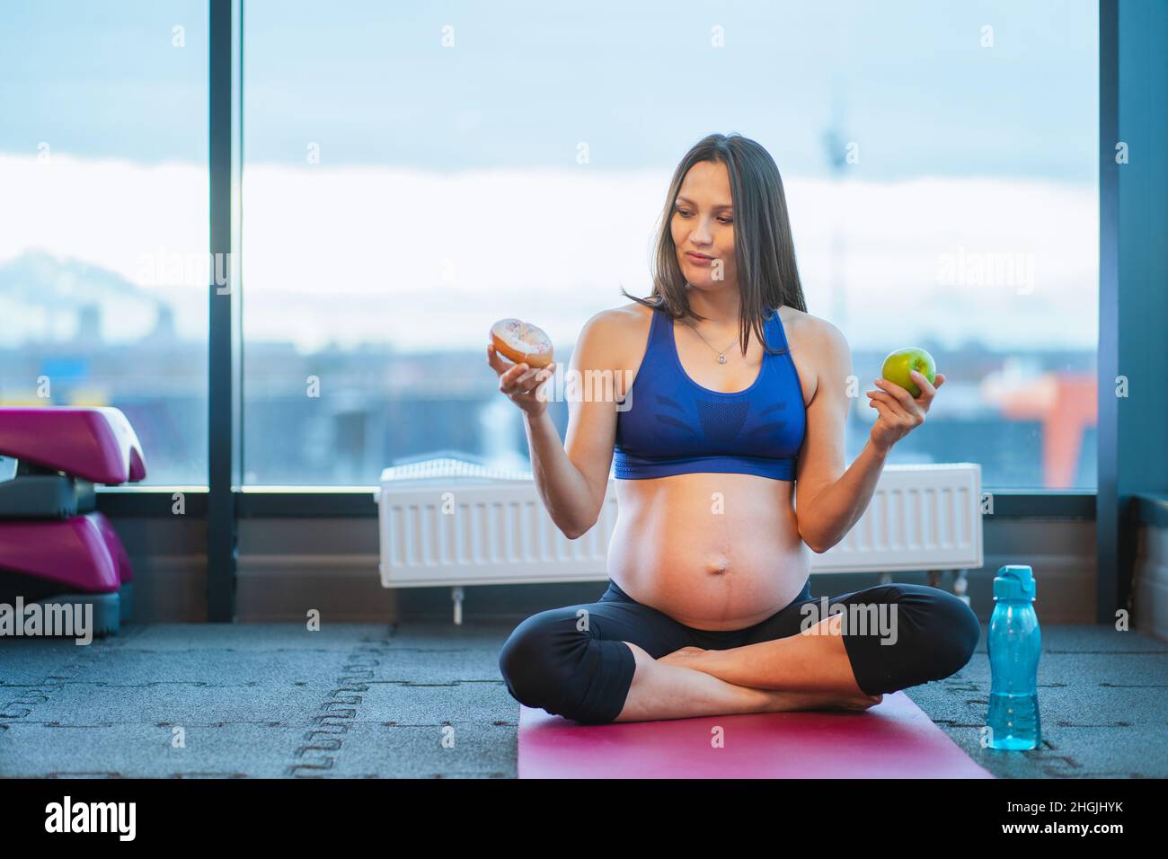 athletic pregnant woman make choice between donut and apple sitting on yoga mat Stock Photo