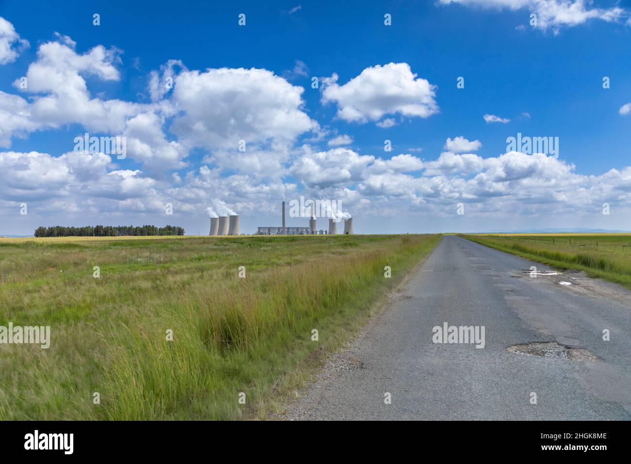 Damaged roadway with large potholes leading into the distance. In the background is a coal fired power plant. Stock Photo
