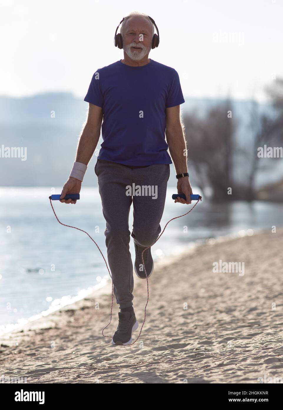 Senior man with beard in sportswear with headphones jumps over a jumping  rope on the beach Stock Photo - Alamy