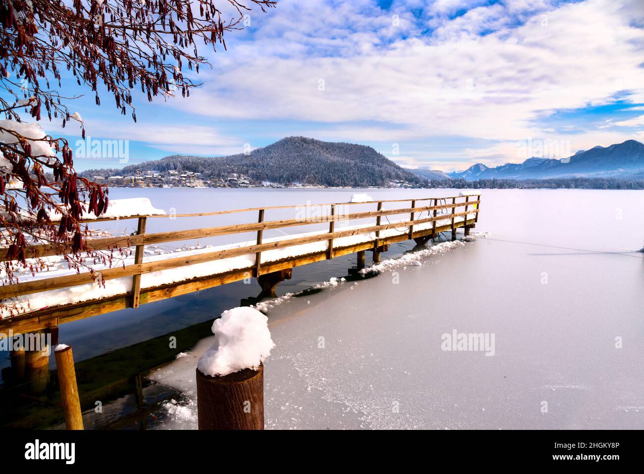 A panoramic view on the Faaker lake in Austrian Alps. The lake is surrounded by high mountains. There is a snow covered pier going into the frozen lak Stock Photo