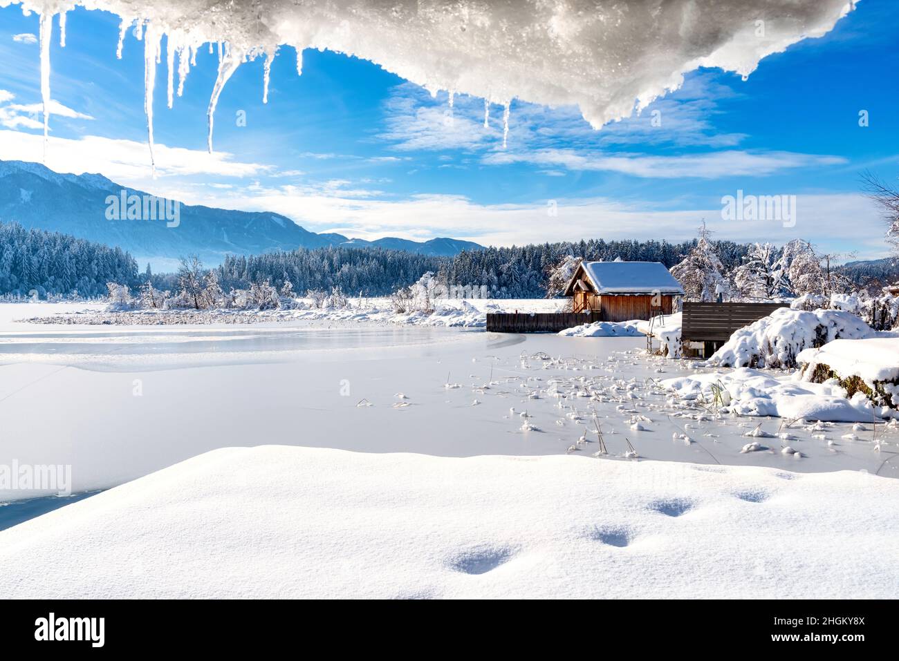 A panoramic view on the Faaker lake in Austrian Alps. The lake is surrounded by high mountains. There is a snow covered pier going into the frozen lak Stock Photo