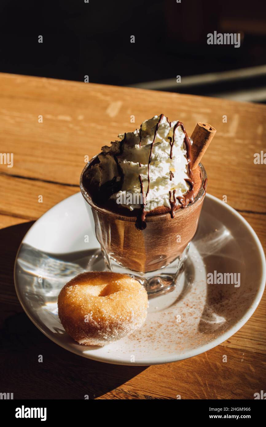 hot drinking chocolate topped with whipped cream, chocolate drizzle, and cinnamon stick on little plate with mini donut Stock Photo