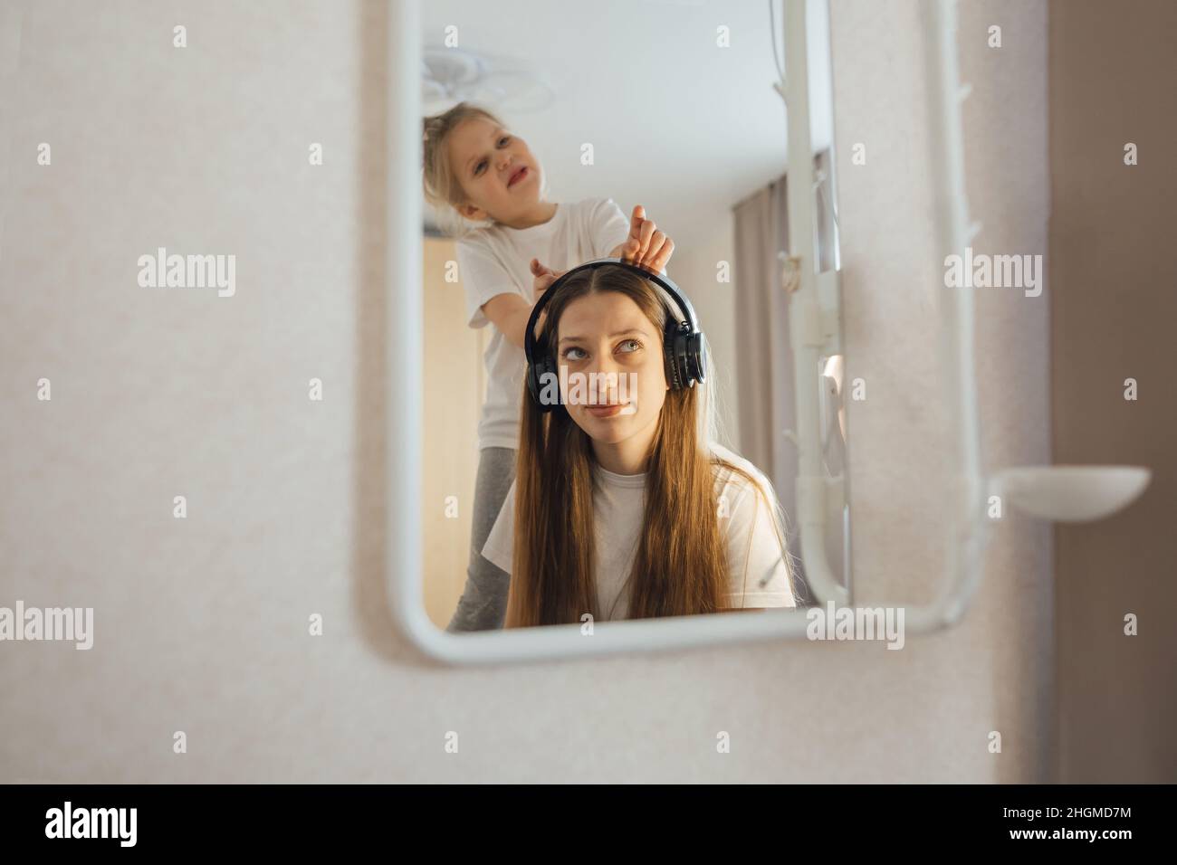 Teenage girl in headphones in front of a mirror, little sister interferes, grimaces. Stock Photo