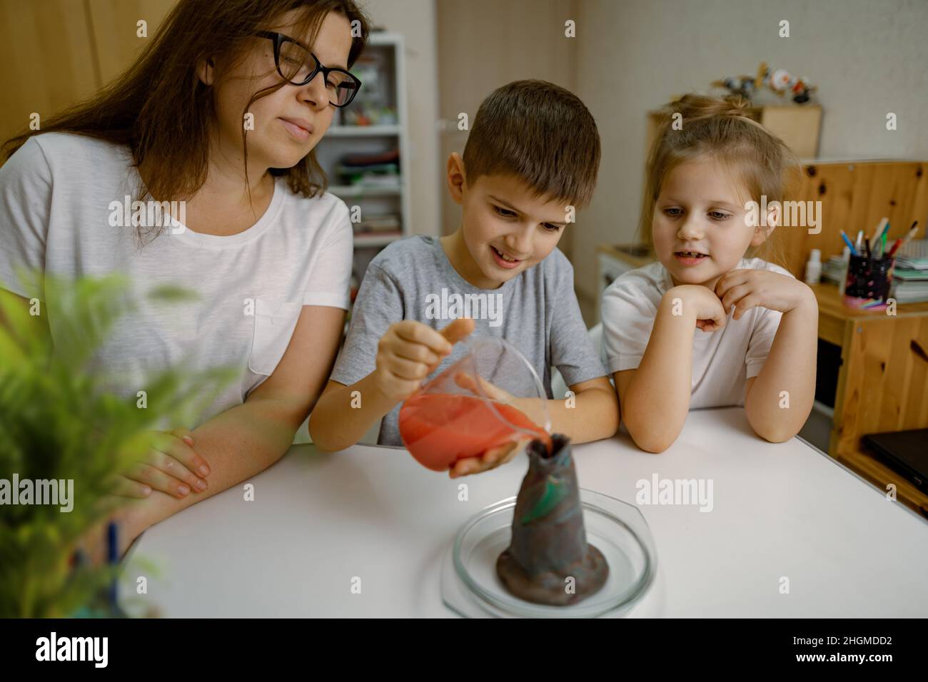 Mom and children at home are conducting an experiment with volcanic eruption. Boy pours liquid into volcanic and rejoices at reaction of eruption. Exp Stock Photo