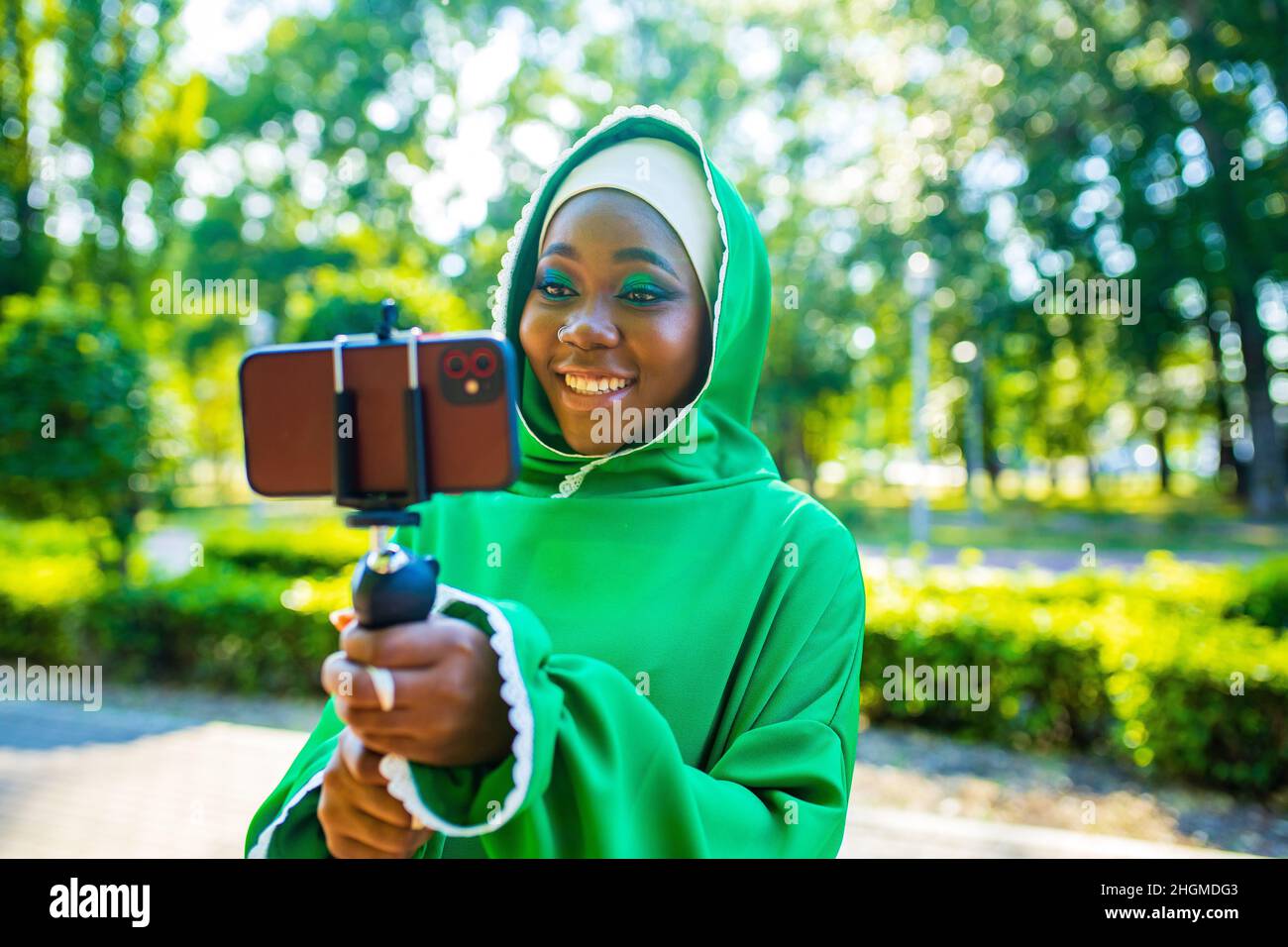 latin hispanic wonan in green muslim hijab with bright make up and nose piercing taking video stream outdoors summer park Stock Photo