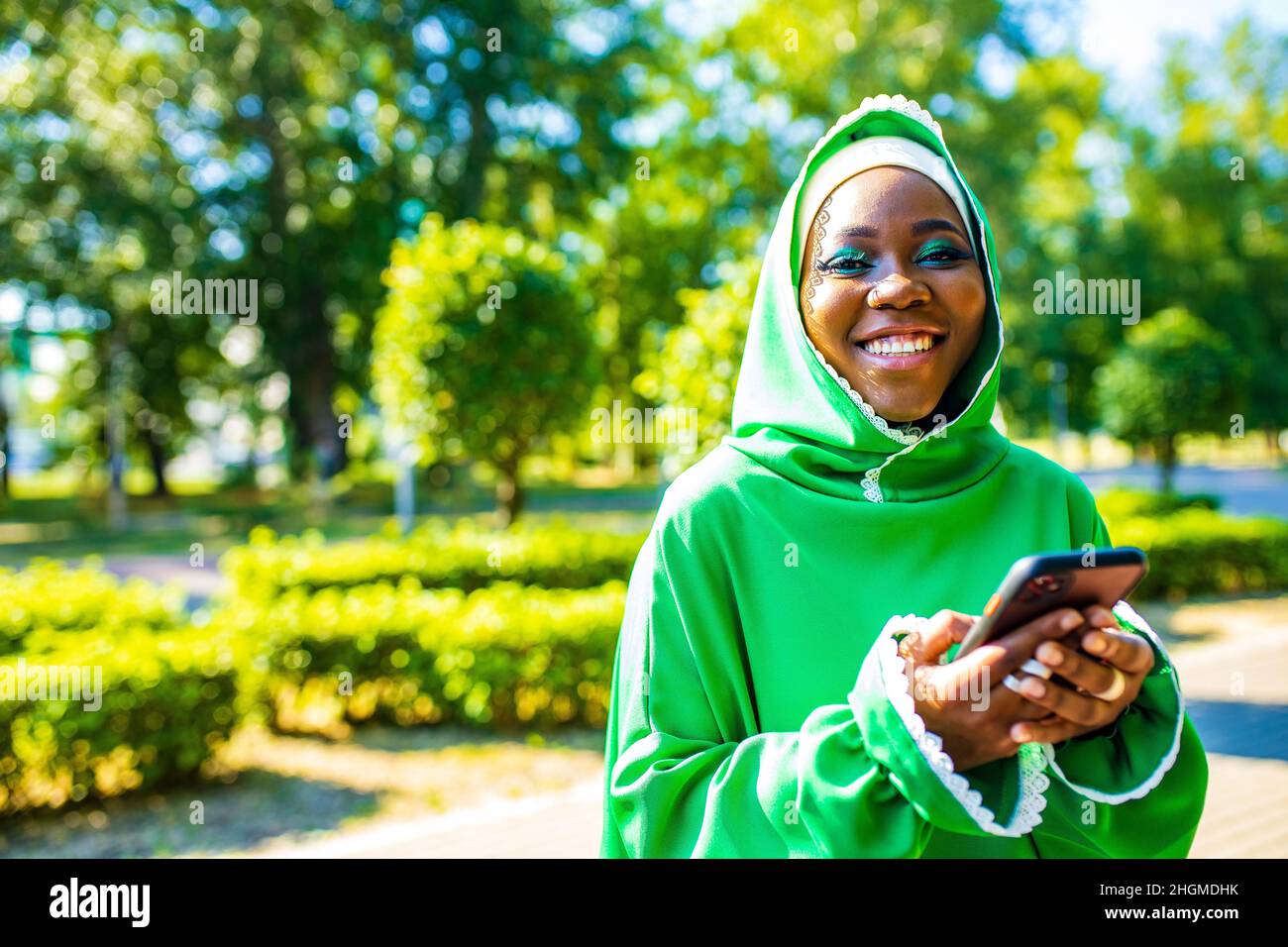 latin hispanic arab woman in green muslim dress with modern bright make up and nose piercing looking at phone screen outdoors Stock Photo
