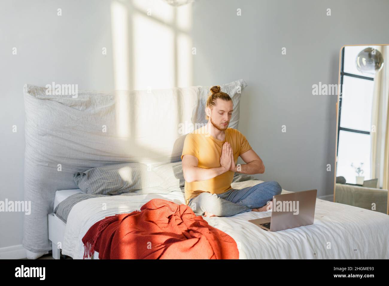 A young man during online meditation on the bed in front of laptop monitor at home. Stock Photo