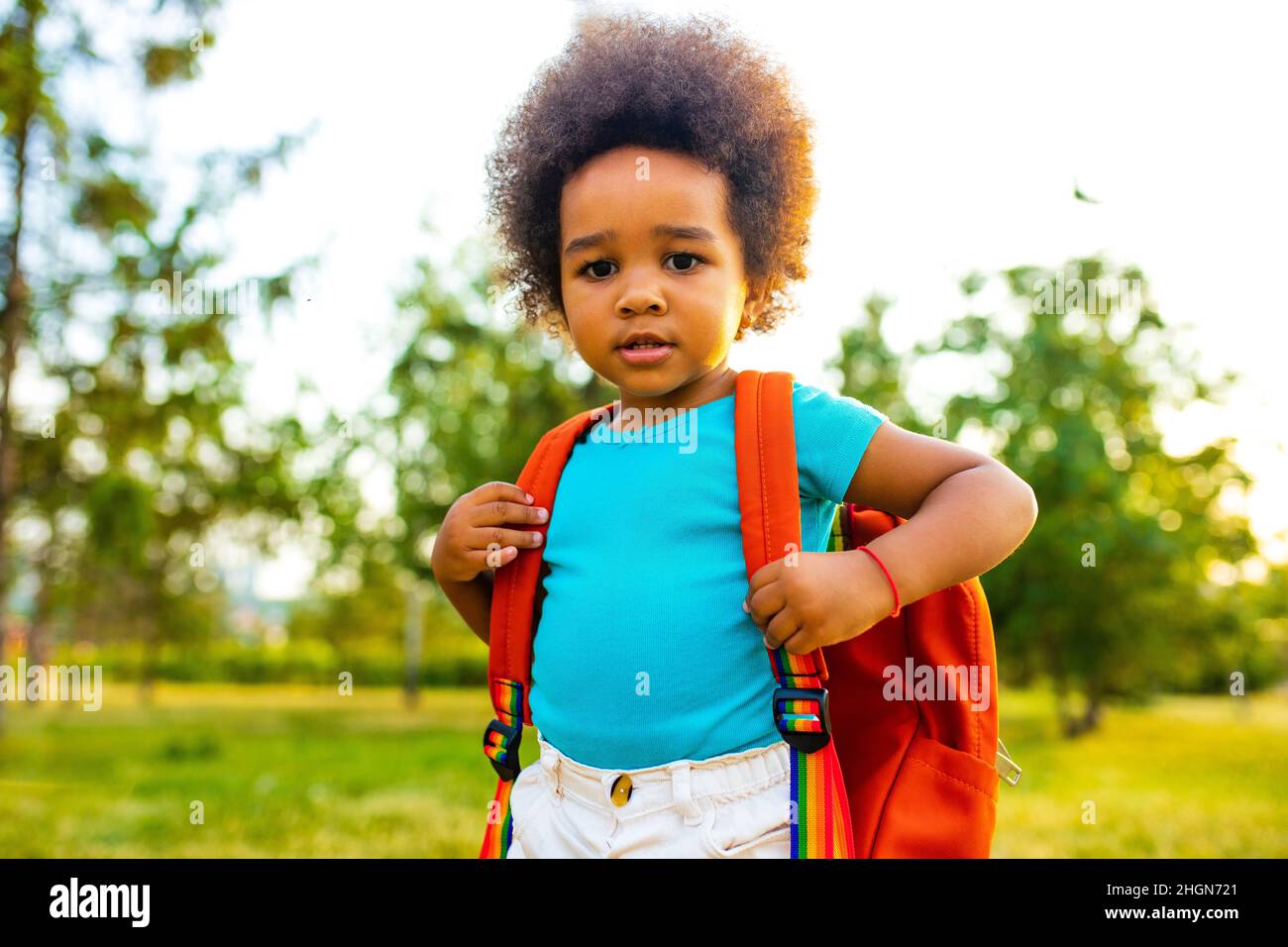 mixed race cute little girl with an orange backpack in blue cotton t-shirt looking at camera outdoors in park Stock Photo