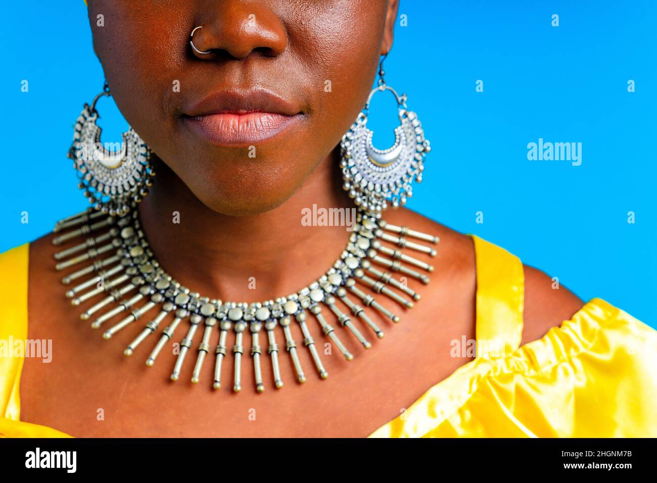 beautiful african woman in yellow silk turban on head and dress with silver neckless and earrings , nose piercing in blue studio background Stock Photo
