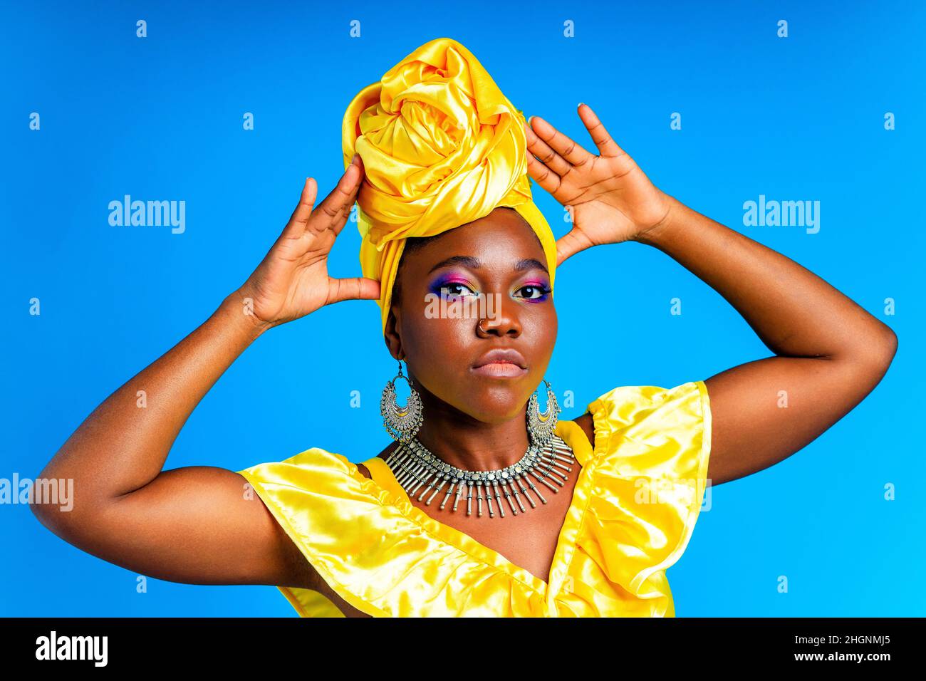beautiful african woman in yellow silk turban on head and dress with silver neckless and earrings , nose piercing in blue studio background Stock Photo