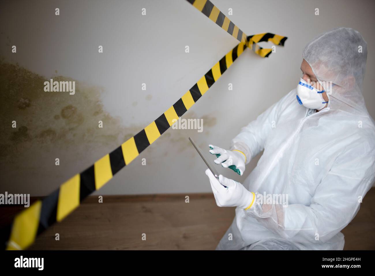 Man with white protective suit and mouth nose mask stands in front of mold on wall and works with tablet behind barrier Stock Photo