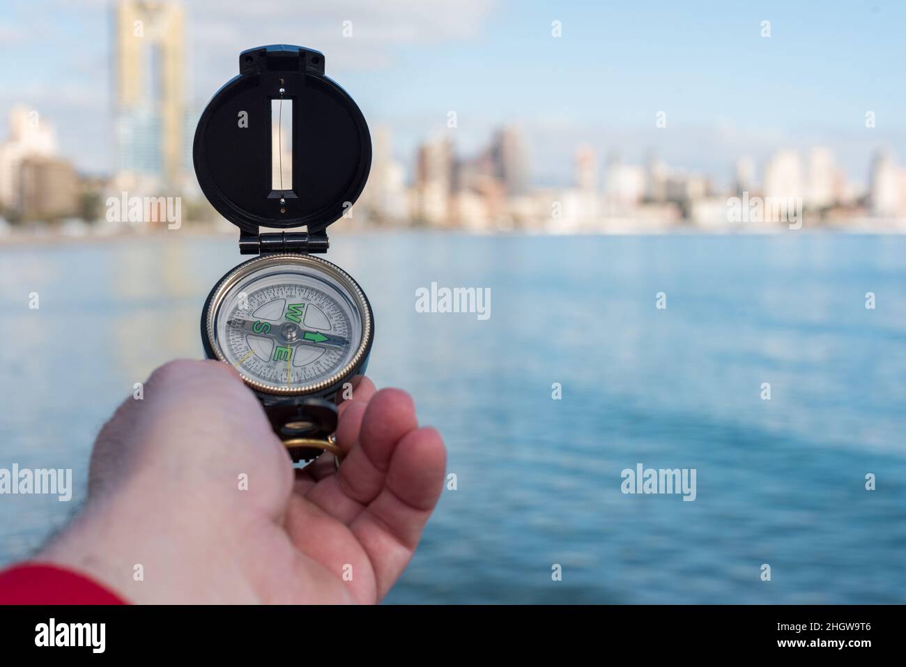 Hand holds a compass over the sea to orient in the city with an out-of-focus city skyline in the background. Stock Photo