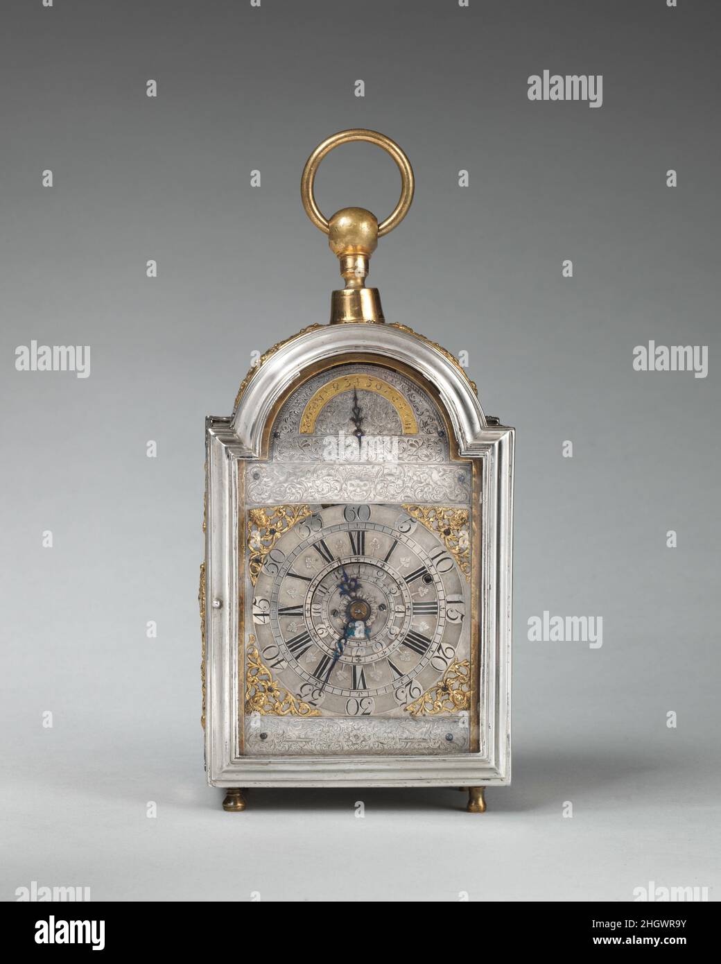 Traveling clock with alarm and calendar ca. 1700–1710 Probably by Joseph Paulet In the late seventeenth and early eighteenth century small clocks adapted for traveling were comparatively rare. This example, in an exquisite case of engraved and repoussé silver, with a champlevé silver dial, belongs to a group of five or six examples, all signed 'Paulet London.' The universal ball joint at the top allows the clock to swing from its suspension ring, cushioning the jolts of motion.. Traveling clock with alarm and calendar. British, London. ca. 1700–1710. Case and dial: silver, partially gilded; br Stock Photo