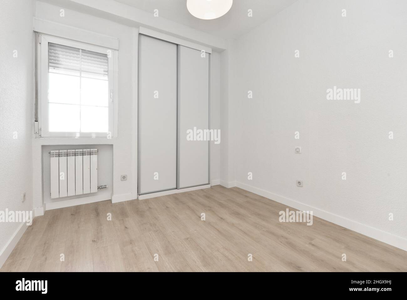 Empty room with light wooden floor, sliding white wooden wardrobe doors and aluminum window above aluminum radiator and ceiling lamp Stock Photo