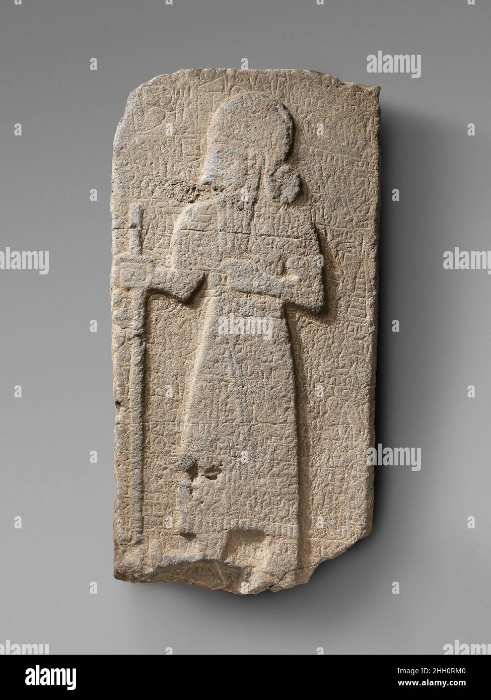 Relief: bearded figure holding staff; hieroglyphic inscription ca. early 1st millennium B.C. Hittite This stele depicts a bearded male figure facing left, surrounded by a shallowly incised inscription divided into seven lines. The figure is shown with attributes characteristic of ruling elites, including the long, fringed garment, the staff he holds in his right hand, his rounded headdress, and his hairstyle, in which the hair is gathered at the nape of the neck in a bunch. The surface of the stele is worn and details are no longer visible in many areas, including the face. The feet are damage Stock Photo