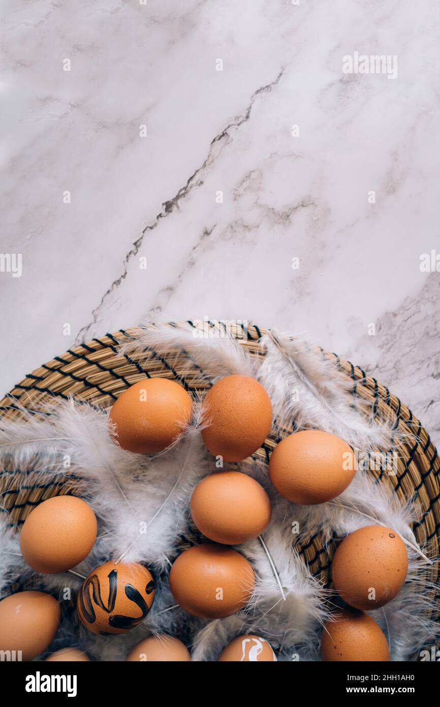 Top view composition of brown chicken eggs and white fluffy birds feathers on round wicker stand on marble table. Easter holiday decorations, flat lay Stock Photo