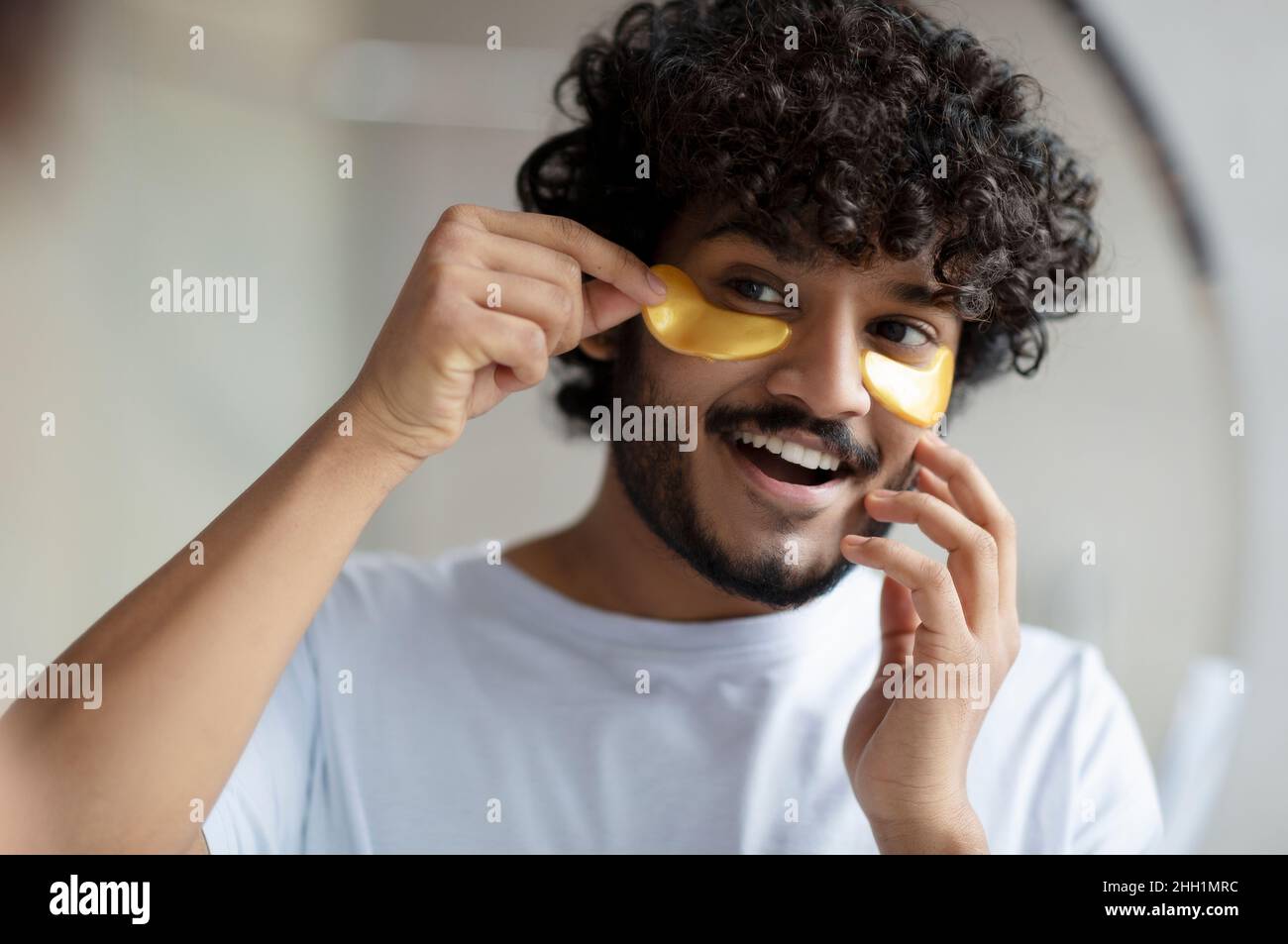 Skin care to reduce eye bags. Cheerful indian man applying gold anti